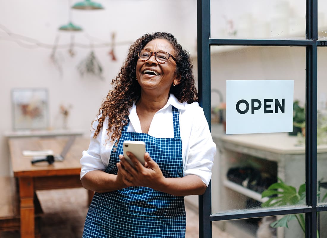 Business Insurance - Cheerful Portrait of a Middle Aged Woman Holding a Phone While Standing Next to the Open Sign on the Front Door of her Shop